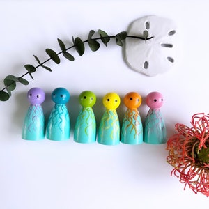 May include: Six colorful wooden peg dolls, each painted with a different color head and a turquoise body with painted tentacles. The dolls are arranged on a white surface with a sprig of greenery, a sand dollar, and a red flower.