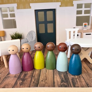 Puede incluir: Siete muñecas de madera de clavija en fila sobre un suelo de madera. Las muñecas están pintadas en diferentes colores: morado, granate, amarillo, verde, azul claro y azul oscuro. Cada muñeca tiene una cabeza marrón con dos puntos negros para los ojos.