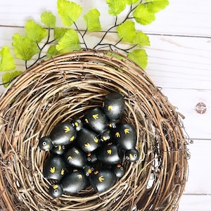 May include: A rustic, woven wreath holds a collection of small, black wooden bird figurines with yellow accents. The birds are nestled within the wreath, which is adorned with green leafy sprigs. The arrangement is set against a white wooden background.