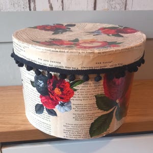 May include: Circular decorative box with a lid, decorated with vintage book pages. It features red and black floral designs and a black pom-pom trim. A unique storage solution.
