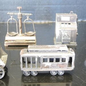 Puede incluir: Una colección de seis figuras de metal en miniatura que incluye un reloj de pie sobre una diligencia, una balanza, una máquina tragamonedas, una rueca, un tranvía de San Francisco y un pozo de agua con cubo y cadena.