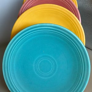 Op de afbeelding: Een stapel van vier ronde keramische borden gerangschikt in een verticale rij. De borden zijn voorzien van een patroon van concentrische ringen op de rand en in het midden. De kleuren van voor naar achter zijn turquoise, geel, oudroze en felgeel.
