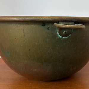 May include: A large, deep, round bowl made of aged brass with a dark, weathered patina. The bowl features a thick, rolled rim and a single metal ring handle attached to the side with a circular plate, showing signs of oxidation and surface wear.