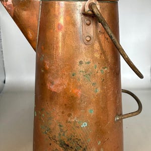 May include: A tall, cylindrical copper coffee pot with a weathered patina, featuring a prominent spout, a domed lid with a small knob, a hinged metal handle attached to the side, and a smaller curved handle on the opposite side of the body.