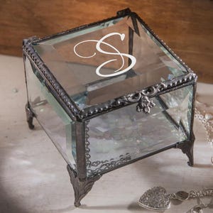 May include: A clear glass jewelry box with a decorative metal frame and legs. The box features a white cursive letter "S" on the lid. The box has a hinged lid and a decorative clasp. The box is empty.