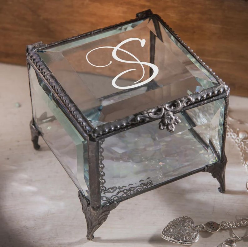 May include: A clear glass jewellery box with a decorative metal frame and legs. The box features a white cursive letter "S" on the lid. The box has a hinged lid and a decorative clasp. The box is empty.