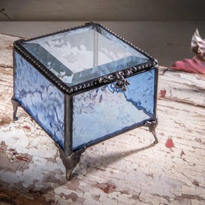 May include: A small, square glass trinket box with a blue and white marbled design. The box has a clear glass lid and sits on four metal feet.