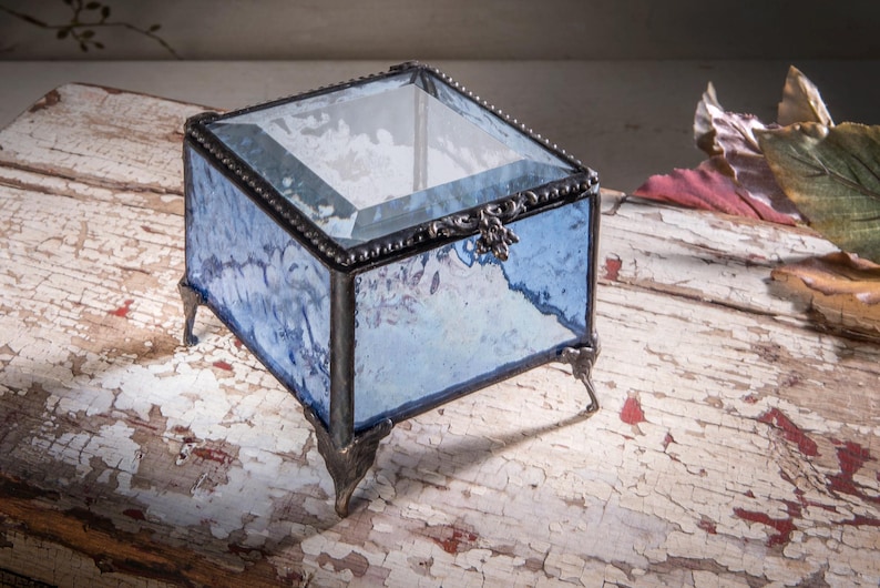 May include: A small, square glass trinket box with a blue and white marbled design. The box has a clear glass lid and sits on four metal feet.