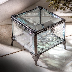 May include: A clear glass ring box with a silver metal frame and ornate detailing. The box has four legs and is sitting on a white surface.