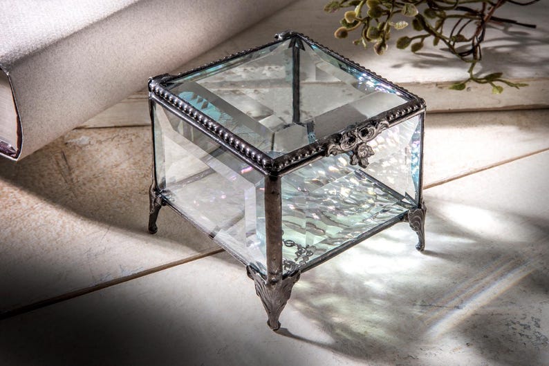 May include: A clear glass ring box with a silver metal frame and ornate detailing. The box has four legs and is sitting on a white surface.