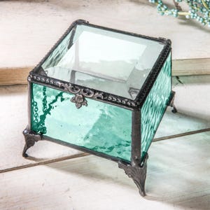 May include: A small, square, teal glass trinket box with a clear glass lid and ornate metal accents. The box has four small feet.
