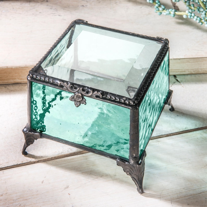May include: A small, square, teal glass trinket box with a clear glass lid and ornate metal accents. The box has four small feet.