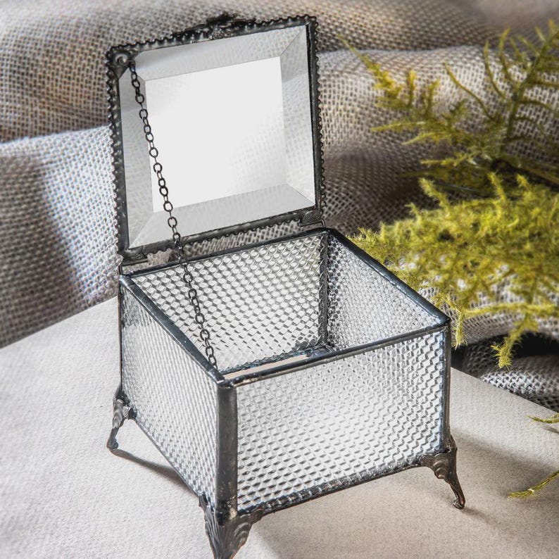 May include: A clear glass trinket box with a hinged lid and a decorative chain. The box has a textured, diamond-patterned design and sits on ornate metal feet. The lid is open, revealing the interior.