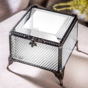 May include: A small, square glass trinket box with a clear, textured glass lid and a black metal frame. The box has four decorative feet and a floral design on the front.