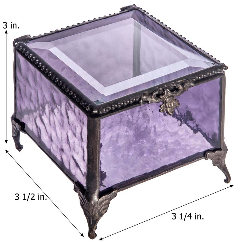 May include: A small, square, purple glass trinket box with a clear glass lid and ornate metal accents. The box has four metal feet and measures 3 1/2 inches wide, 3 1/4 inches deep, and 3 inches tall.