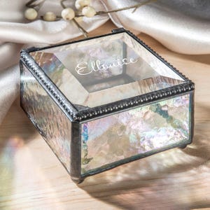 May include: Iridescent glass jewelry box with a silver-colored metal frame. The box has a hinged lid and is inscribed with the name "Ellouise". The glass has a textured, crackled appearance, and the box sits on small feet.