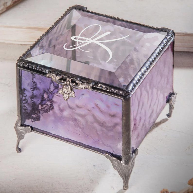 May include: A decorative glass box with a purple tint, featuring a hinged lid and ornate metal detailing. The lid displays a white letter "K". The box is supported by decorative metal feet.