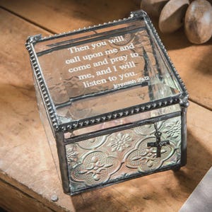 May include: A clear glass box with a decorative floral pattern on the front and sides. The lid has an etched quote from Jeremiah 29:12. A small cross charm hangs from the front. The box is trimmed with a dark metal.