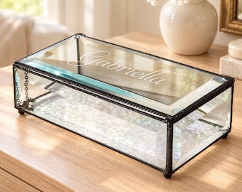 May include: A rectangular glass keepsake box with a dark metal frame and a hinged lid. The top glass panel features the engraved name Gabriella in a cursive script. The base of the box has a textured, iridescent finish, and the structure rests on four small, rounded metal feet.