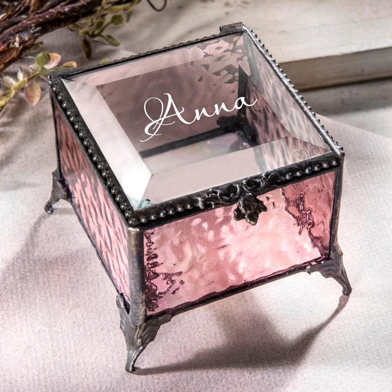 May include: A small, square, pink glass trinket box with a clear glass lid. The box has a decorative metal trim and four metal feet. The lid is engraved with the name "Anna".