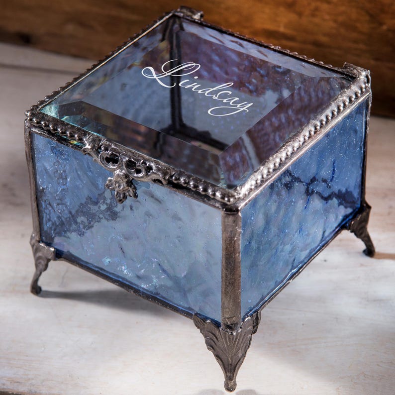 May include: A small, square, blue glass trinket box with a silver metal frame and feet. The top of the box is clear glass and has the name "Lindsay" engraved on it.
