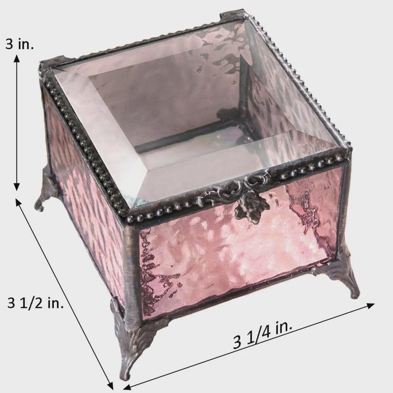 May include: A decorative pink glass box with a hinged lid, measuring 7.6 cm high, 8.9 cm long, and 8.3 cm wide. The box has a metal frame and ornate feet, perfect for storing jewelry or small keepsakes.