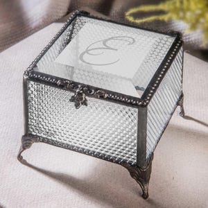 May include: A clear glass jewellery box with a decorative metal frame and feet. The box has a textured diamond pattern on the sides and a hinged lid. The lid features a printed letter "E".