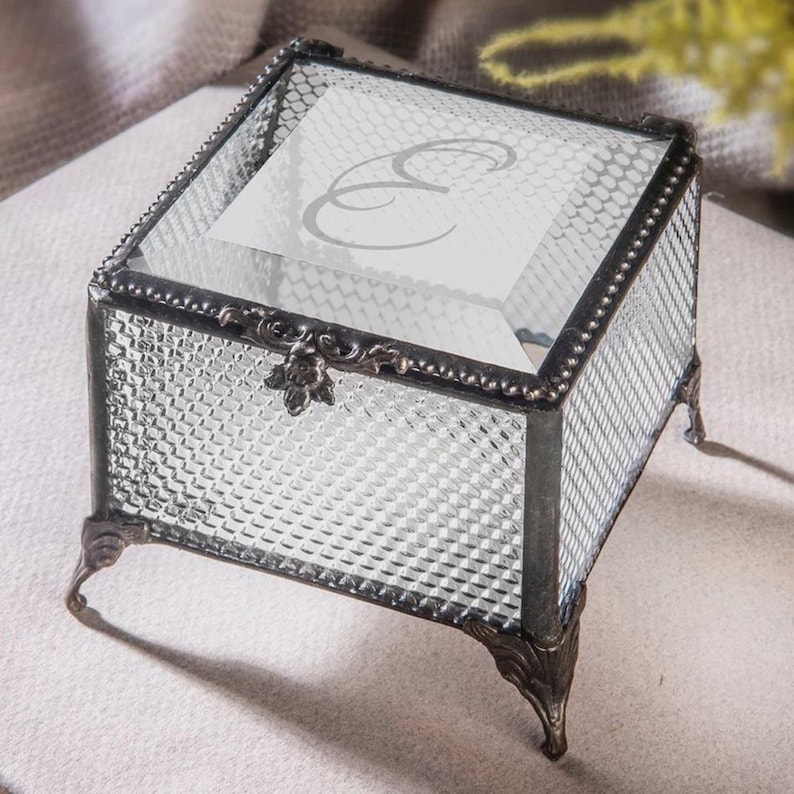 May include: A clear glass jewellery box with a decorative metal frame and feet. The box has a textured diamond pattern on the sides and a hinged lid. The lid features a printed letter "E".