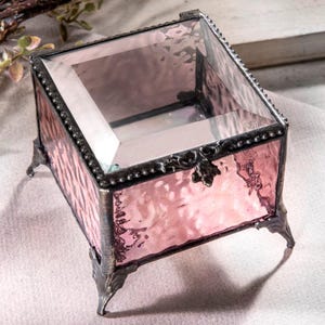 May include: A small, square, pink glass trinket box with a clear glass lid and ornate metal accents. The box has four small feet and a hinged lid.