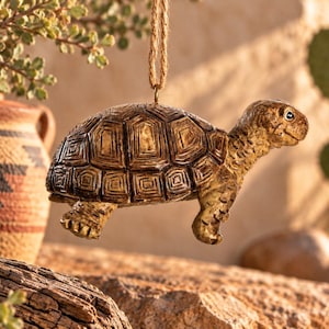 May include: A hanging tortoise ornament featuring a textured brown shell with detailed geometric scutes and a realistic head. The figure is suspended by a thin jute twine loop attached to a small metal eyelet on the top of the shell.
