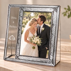 May include: A rectangular silver-toned metal picture frame with an embossed floral border. The frame features a side panel etched with the text Olivia & Matthew, a decorative letter S, and the date 06.15.26, holding a wedding photograph.
