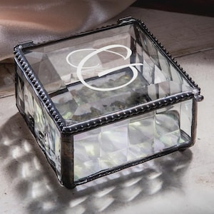 May include: A clear glass keepsake box with a decorative metal frame and a white letter "C" on the lid. The box has a geometric design and is suitable for storing small items. The box is on a neutral surface.