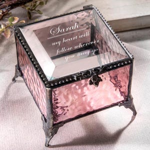 May include: A small, square, pink glass trinket box with a clear glass lid. The lid is engraved with the text "Sarah my heart will follow wherever you may go". The box has a decorative metal base with four feet.