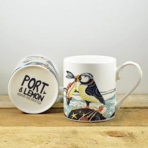 May include: Two white ceramic mugs. One mug is upright, displaying a colorful illustration of a puffin bird with a fish in its beak. The other mug is upside down, revealing the text "PORT & LEMON HAND DECORATED". The mugs are on a wooden surface.