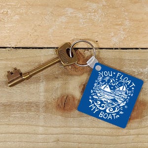 Op de afbeelding: Een blauwe vierkante sleutelhanger met de tekst "You Float My Boat" in witte letters. De sleutelhanger heeft een witte illustratie van twee zeilboten op een golf met sterren en vissen. De sleutelhanger is bevestigd aan een gouden sleutel.