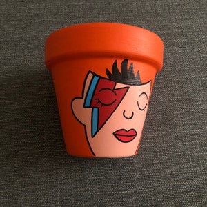 May include: A terracotta flower pot painted with a portrait of David Bowie. The portrait is in a cartoon style and features Bowie's signature lightning bolt makeup. The pot is a light orange colour.