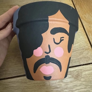 May include: A hand-painted terracotta plant pot with a stylized face design. The pot features black hair, a mustache, pink cheeks and lips, and a light purple rim. The face is painted on a brown background. The design is artistic.