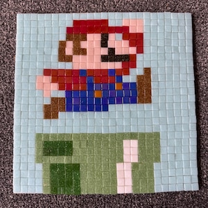 May include: A mosaic tile artwork depicting a pixelated image of Mario jumping over a green pipe. The artwork uses red, blue, brown, and light blue tiles to recreate the iconic video game character and scene.