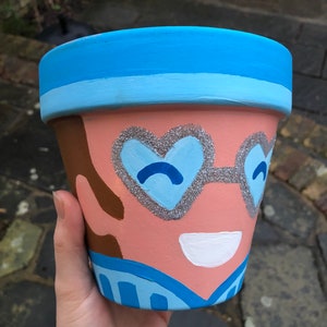 May include: A hand-painted terracotta flower pot with a whimsical face design. The pot has a light blue rim, a peach-coloured face, and heart-shaped glitter glasses. Brown hair and a blue and white striped shirt complete the design.