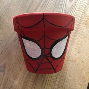 May include: A red terracotta plant pot decorated with a Spider-Man mask design. The pot has a red base with black web lines and white eye shapes. Suitable for indoor or outdoor use, this pot is ideal for plants.