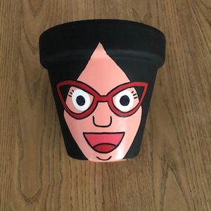 May include: A hand-painted terracotta flower pot featuring a cartoon face. The face has black hair, red glasses, a pink face, and a red smile.