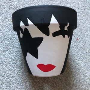 May include: A black and white painted terracotta flower pot with a stylised face and red lips. The face is painted to resemble a rock star with a star on the forehead.
