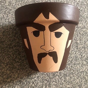 May include: A brown terracotta flower pot painted with a cartoon face. The face has black eyes, eyebrows, and a moustache.