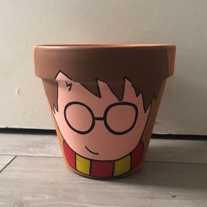May include: A terracotta flower pot painted with a cartoon illustration of Harry Potter wearing glasses and a red and yellow scarf.