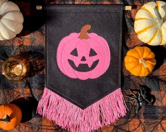 May include: A black felt Halloween banner with a pink pumpkin design and pink fringe. The banner is displayed with small pumpkins, a candle holder, and a plastic spider on a plaid background.