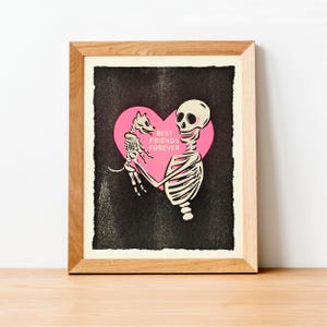 Puede incluir: Una impresión enmarcada que presenta un esqueleto sosteniendo un perro esqueleto dentro de un corazón rosa. Las palabras "BEST FRIENDS FOREVER" están impresas en el corazón. El fondo es negro con un efecto texturizado, y el marco es de madera clara.