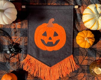 May include: A black Halloween banner with an orange pumpkin design and fringe. The banner is hanging on a wooden dowel. The image also includes pumpkins, a spider, and a candle.