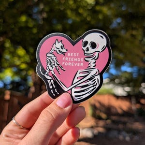 Peut inclure: Autocollant rose en forme de c&oelig;ur avec des illustrations de squelettes d'un chien et d'une personne qui s'embrassent. Le texte "BEST FRIENDS FOREVER" est imprim&eacute; en blanc. L'autocollant a une bordure noire.