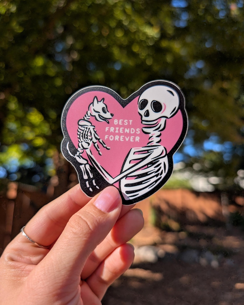 Peut inclure: Autocollant rose en forme de c&oelig;ur avec des illustrations de squelettes d'un chien et d'une personne qui s'embrassent. Le texte "BEST FRIENDS FOREVER" est imprim&eacute; en blanc. L'autocollant a une bordure noire.