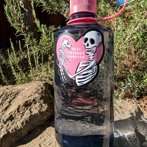 Peut inclure: Une bouteille d'eau transparente avec un bouchon et une poign&eacute;e roses. Un autocollant rose en forme de c&oelig;ur repr&eacute;sente un squelette tenant un chien avec le texte "BEST FRIENDS FOREVER". La bouteille est remplie d'un liquide fonc&eacute;.
